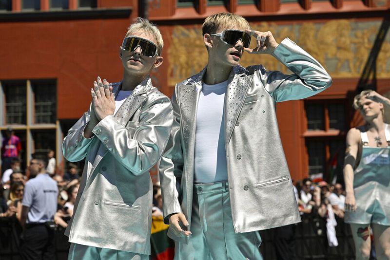 Dos personas con conjuntos plateados y turquesa, gafas de sol y chaquetas brillantes posan en una alfombra.  Fondo con más gente y un edificio.