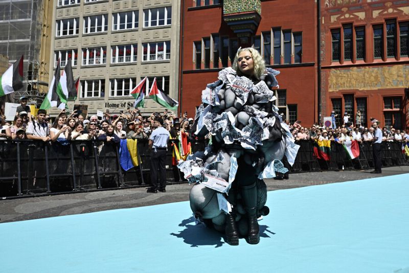 Mujer con vestido de papel arrugado posa en alfombra turquesa; fondo con multitud, banderas y edificios.  Posible evento público.