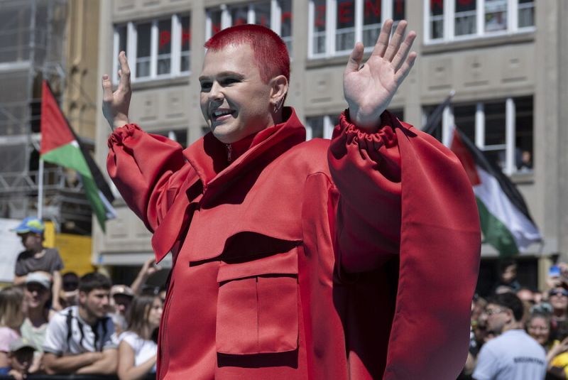 Persona con cabello rojo y atuendo rojo amplio saluda a la multitud en una alfombra turquesa.  Se aprecian mangas largas y un bolsillo en la prenda.