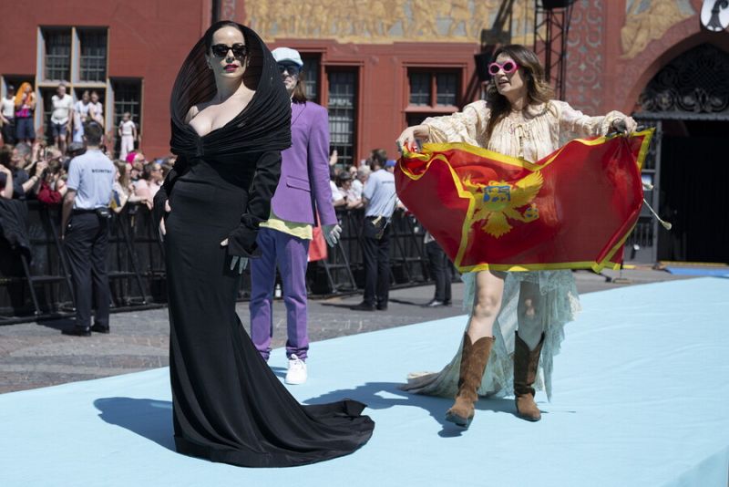 Dos mujeres con estilos diferentes posan en una alfombra turquesa; una con vestido negro y gafas oscuras, la otra con vestido bohemio, botas marrones, gafas rosas y la bandera de Montenegro.