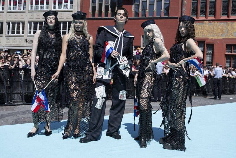 Grupo de cinco personas con atuendos negros, gorros de marinero y detalles de red, posando en una alfombra.  Las mujeres portan pequeñas banderas.