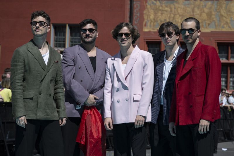 Cinco hombres con americanas de colores variados y gafas (algunos graduadas) posan; uno lleva una tela roja.  Posible alfombra roja de evento.