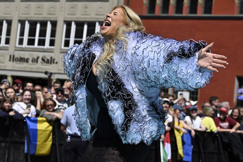 Mujer rubia con chaqueta azul claro y expresión alegre, rodeada de público en una alfombra turquesa. Se observan banderas, entre ellas la de Ucrania.
