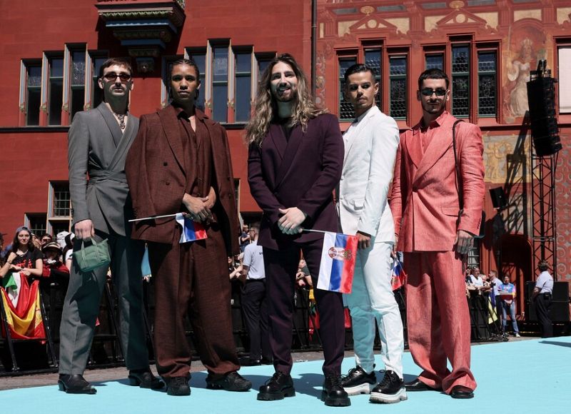 Grupo de cinco personas en trajes coloridos con banderas serbias, posando para una foto en una plaza.  Público visible al fondo.