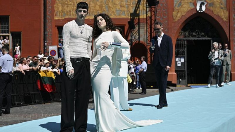 Abor y Tynna, en la Alfombra Turquesa, lucen atuendos elegantes: él con camiseta clara y pantalones negros, ella con un vestido blanco.  Se observa público y un edificio al fondo.