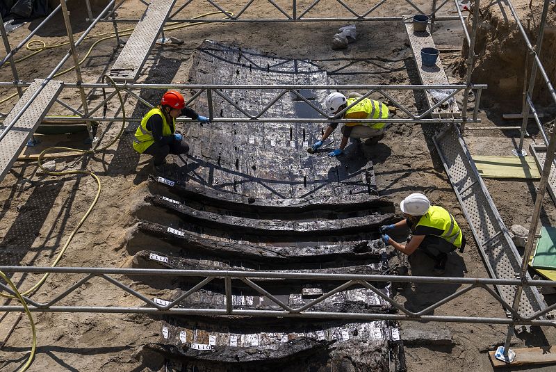 Arqueólogos excavan una estructura de madera numerada, posiblemente un barco, usando andamios y herramientas.  El suelo es de tierra.
