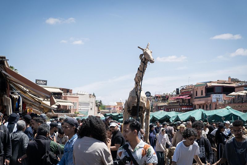 Marioneta gigante de jirafa en un mercado marroquí, rodeada de gente que la observa y la fotografía durante un desfile.