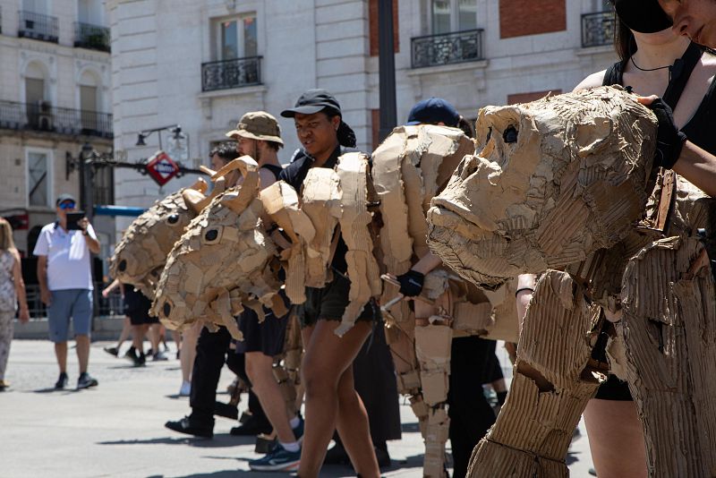 Marionetas gigantes de cartón con forma de animales, posiblemente toros y primates, interactúan con la gente en una plaza soleada.  Aspecto artesanal.