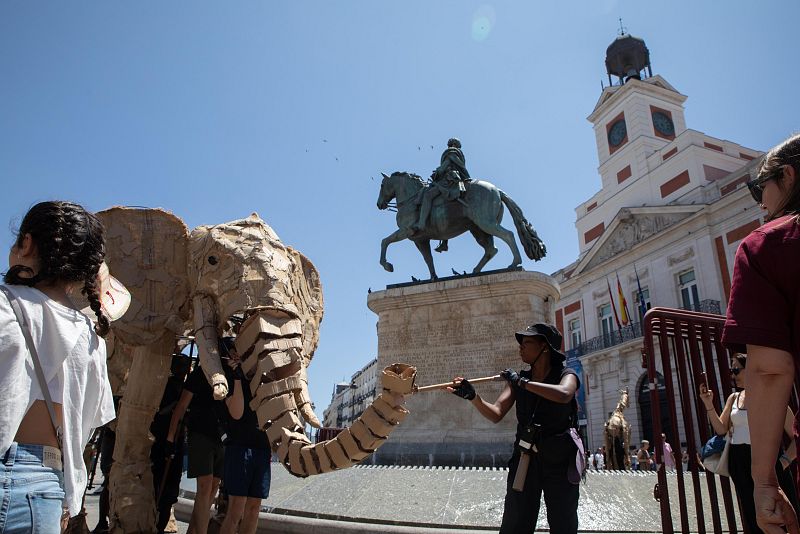 Marioneta gigante de elefante en la Puerta del Sol, manipulada por una persona con vara. Niña observa la escena, fondo con estatua ecuestre y Casa de Correos.