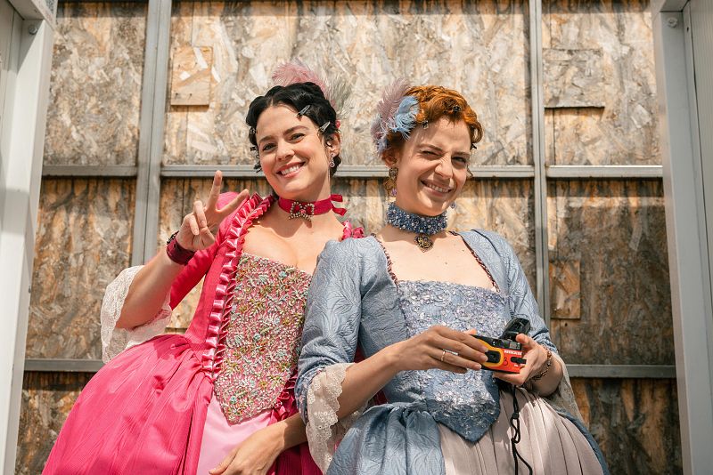 Dos mujeres con vestidos de época y accesorios modernos posan: una con vestido rosa y símbolo de paz, la otra con vestido azul y cámara instantánea. Fondo de madera.