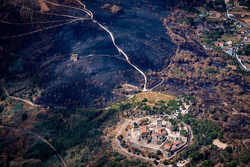 Vista aérea de un castillo en una colina, rodeado de una zona quemada tras un incendio forestal.  El castillo parece intacto, con caminos visibles en el paisaje desolado.