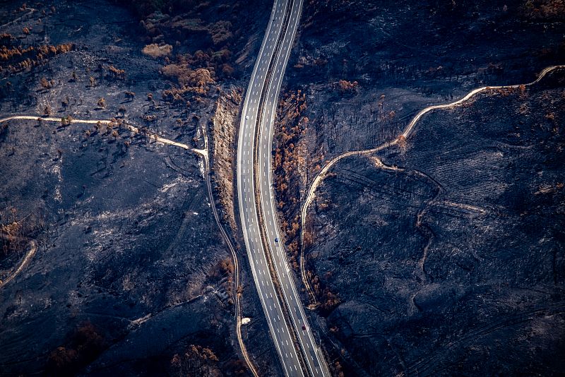 Autovía tras incendio forestal: contraste entre el asfalto intacto y el terreno quemado circundante.  Se ven vehículos en circulación.