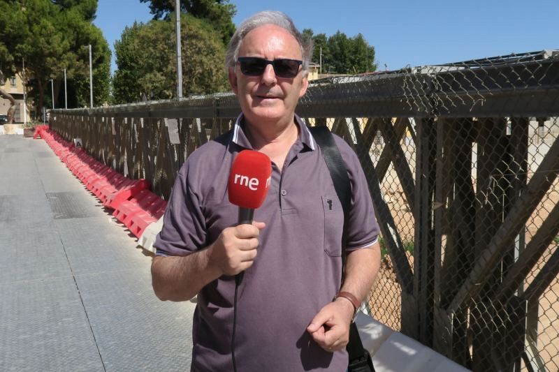 Un hombre con gafas de sol y camisa morada, posiblemente entrevistado, sostiene un micrófono con el logo de una emisora de radio. Se encuentra en un lugar elevado con barandillas metálicas y barreras de seguridad.