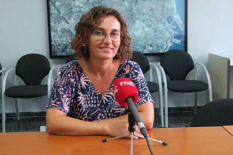 Carmen Zornoza, con gafas y blusa floral, está sentada frente a un micrófono rojo con el logo de una emisora de radio. Al fondo, se aprecian sillas y un cuadro.