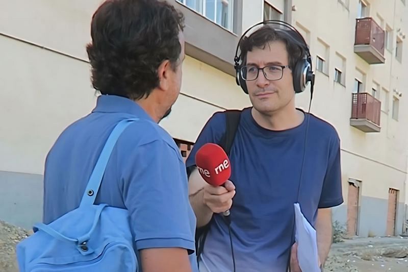 Álvaro Soto, con micrófono y auriculares, realiza una grabación en exteriores. La persona entrevistada, con camisa y mochila azules, está frente a él. El entorno muestra edificios y una zona en obras.