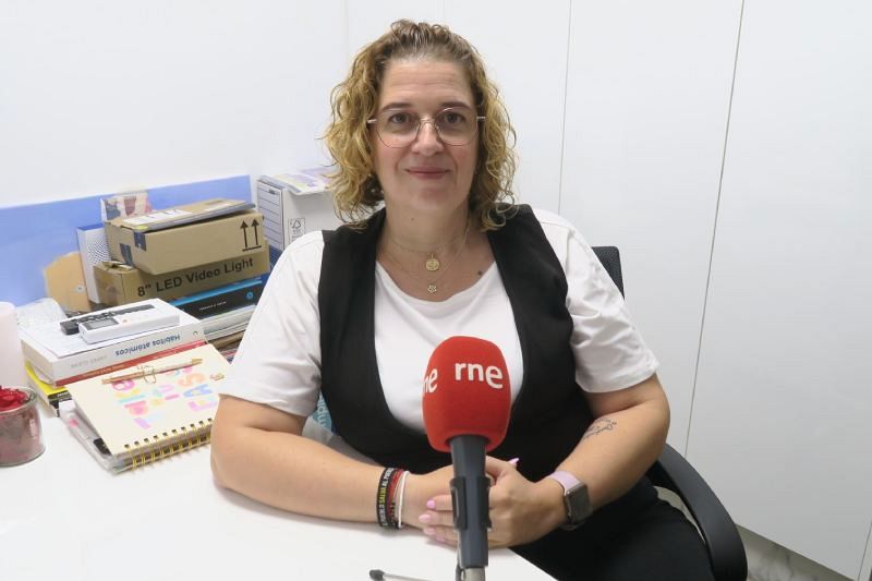 Una mujer con gafas y cabello rizado, vestida con una camiseta y chaleco, está sentada frente a un escritorio con un micrófono rojo y otros objetos.