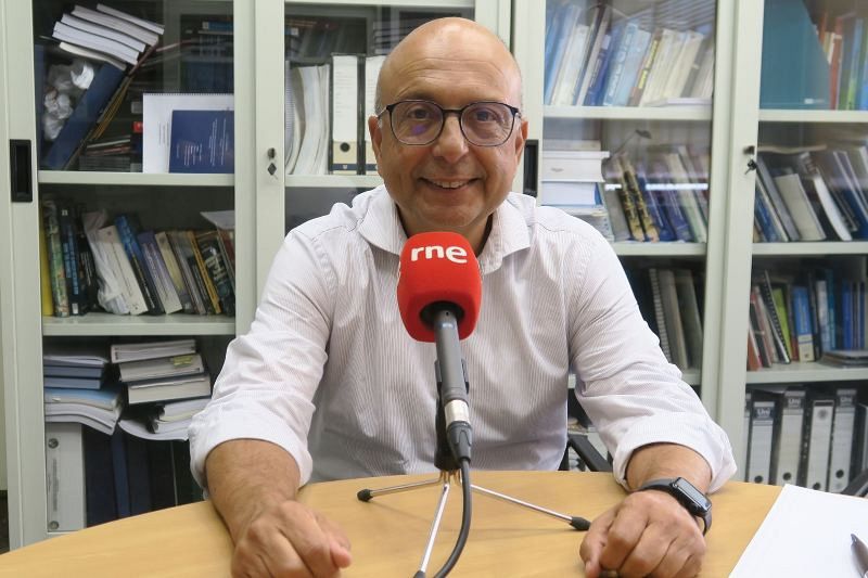 Ignacio Escuder, con gafas y camisa blanca, sonríe a la cámara frente a un micrófono con el logo de una emisora de radio. Se encuentra en un estudio con estanterías llenas de libros.