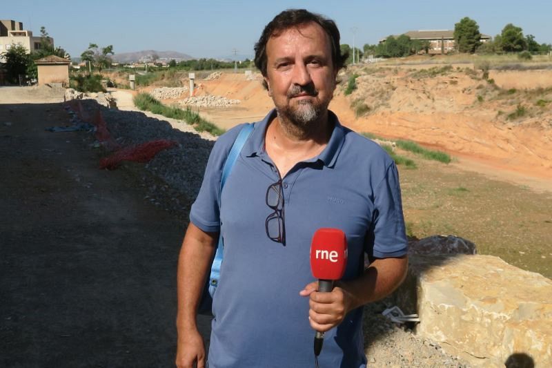 Un hombre de mediana edad, con camisa azul y gafas, sostiene un micrófono con el logo de RNE, en un terreno en obras.