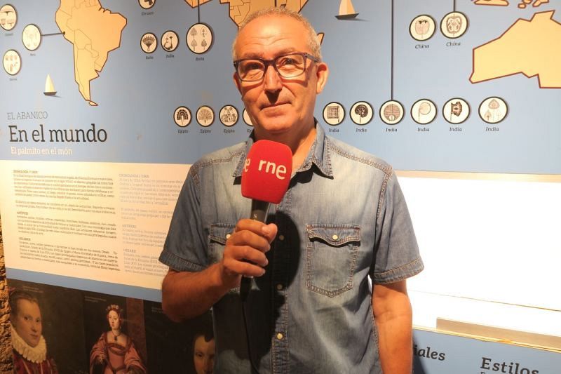 Francesc Martínez Sanchis, con gafas y camisa vaquera, sostiene un micrófono de RNE frente a un mapa mundial con ilustraciones de abanicos y textos en diferentes idiomas, incluyendo nombres de países.