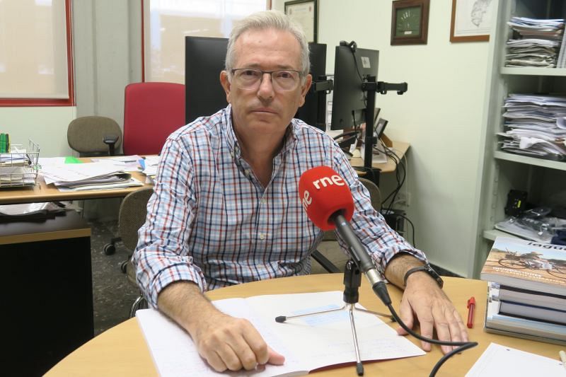 Félix Francés, con camisa de cuadros y gafas, está sentado frente a un micrófono rojo con el logo de una emisora de radio. La mesa frente a él está llena de documentos y un bolígrafo, con monitores y una estantería al fondo.