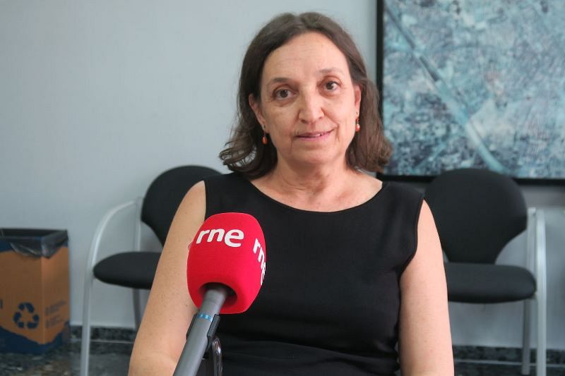 María José López, profesora, aparece frente a un micrófono con el logo de una emisora de radio. Viste un vestido negro y pendientes rojos, con una pintura abstracta y un cubo de reciclaje como fondo.
