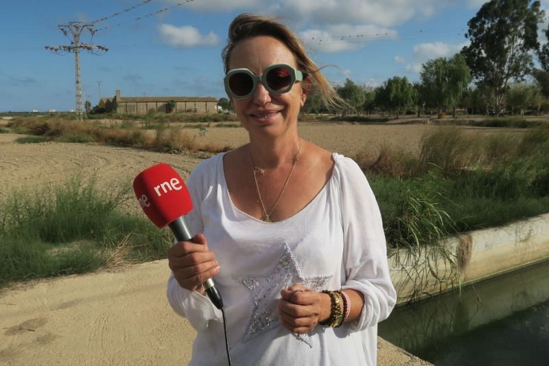 Una mujer con gafas de sol y blusa blanca, sosteniendo un micrófono rojo con un logo, posa frente a un paisaje rural con campos y vegetación, con el cielo parcialmente nublado.