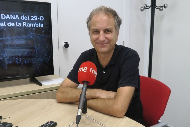 Nacho Payá-Zaforteza, profesor, está sentado frente a un micrófono rojo en un estudio de radio, posiblemente de RNE. Viste una camisa negra y al fondo se observa una pantalla con información.