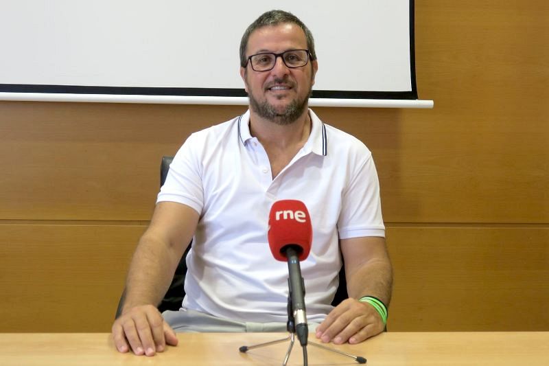Un hombre con barba y gafas, identificado como Miguel Ángel Eguíbar, está sentado frente a una mesa con un micrófono rojo. Viste una camisa polo blanca y lleva una pulsera verde.