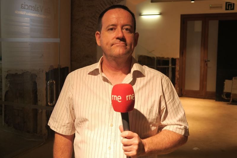 Salvador Collado, con una camisa a rayas, sostiene un micrófono rojo con el logo de una emisora de radio. Se encuentra en un interior con una puerta de madera y una inscripción en la pared.