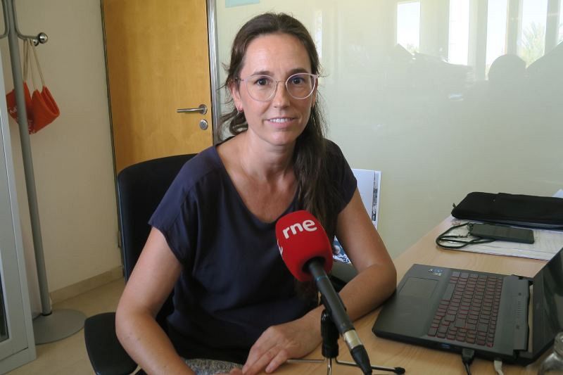 En la imagen, una mujer con gafas y camiseta azul está sentada frente a un escritorio con un micrófono rojo, un portátil, una funda y un teléfono. Se observa una pared blanca con una puerta y un perchero.