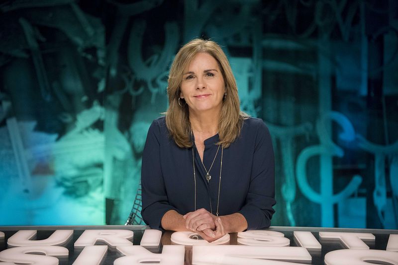 Una mujer de mediana edad, con cabello castaño claro y una blusa azul oscuro, mira directamente a la cámara. Sus manos están entrelazadas sobre una superficie blanca con letras en relieve, con un fondo abstracto en tonos azules y verdes.