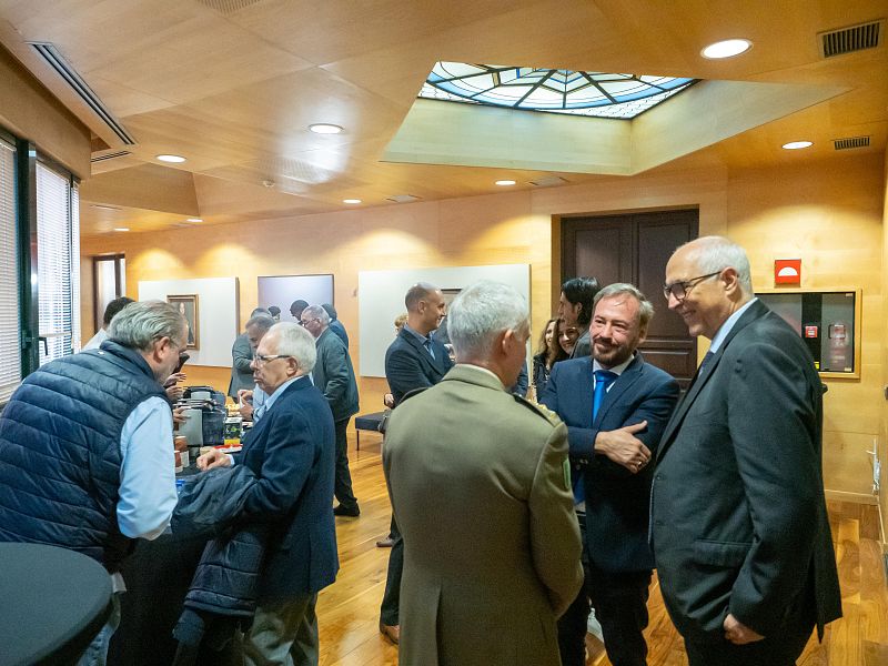 En un evento social, dos hombres vestidos con trajes, uno azul y otro oscuro con gafas, conversan animadamente en un salón con paredes de madera y un tragaluz.
