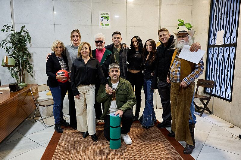 Un grupo de personas posa en un interior, posiblemente un estudio. Se observa una mesa con objetos, una ventana con barrotes y un cartel de reciclaje. La escena transmite una sensación de cercanía entre los individuos.