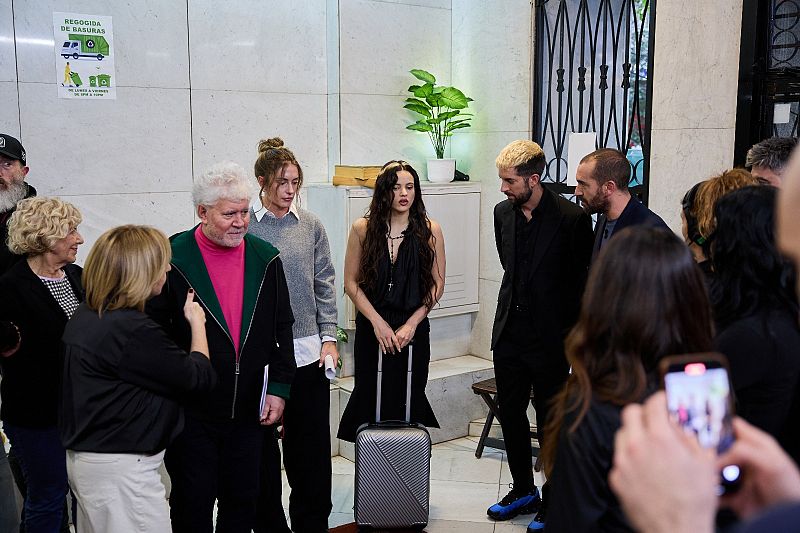 Rosalía, con un vestido negro y un collar, posa con una maleta plateada junto a un hombre con una chaqueta negra y detalles verdes. El entorno es un espacio interior con paredes de mármol, donde otras personas toman fotos.
