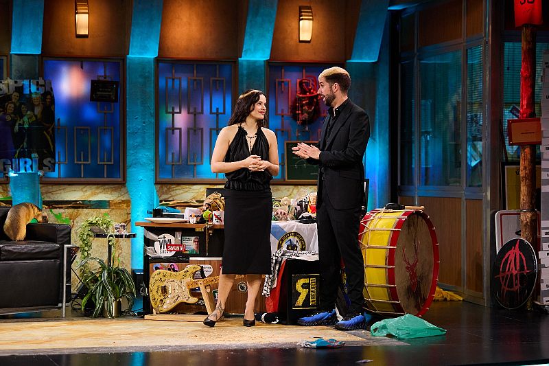 En un plató televisivo, una mujer vestida de negro y un hombre con traje negro y pelo rubio posan juntos. El escenario incluye un sofá, instrumentos musicales y un cartel.