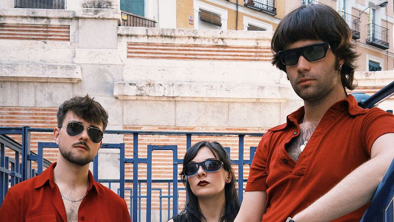 Tres personas posan en un entorno urbano. Un hombre con camisa roja y gafas de sol está a la izquierda, una mujer con top negro y gafas oscuras en el centro, y otro hombre con camisa roja y gafas de sol a la derecha, apoyado en una barandilla.