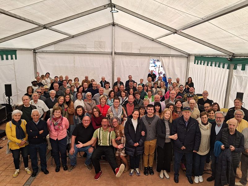 Un evento concurrido se celebra bajo una carpa blanca, con una multitud de personas de diversas edades. La mayoría están de pie, con algunos sentados, y la vestimenta es casual, con algunos detalles coloridos.