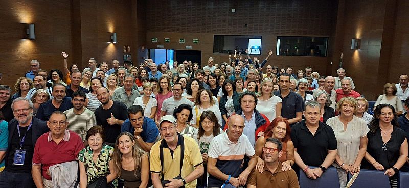 Un grupo numeroso de personas, vestidas de manera informal, posan sonrientes para una foto en un espacio amplio, posiblemente un auditorio, con un ambiente festivo.