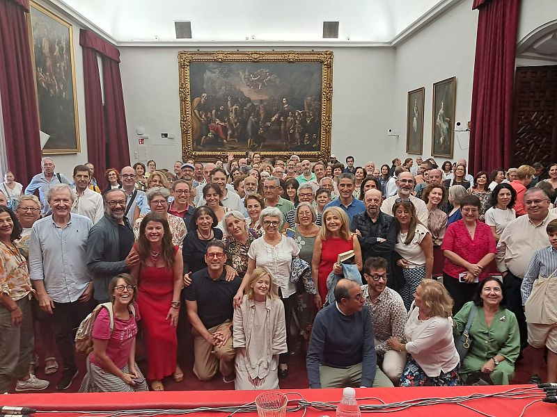 Un grupo numeroso posa en una sala con techos altos, frente a un cuadro clásico y cortinas rojas. La mayoría están de pie, con algunos sentados o arrodillados, todos sonriendo a la cámara. En primer plano, una mesa con mantel rojo.
