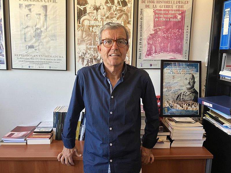 Julián Chaves, con gafas y camisa azul, se encuentra frente a una pared con carteles sobre la Guerra Civil Española, incluyendo uno sobre la represión en Extremadura y otro sobre la memoria histórica de 1936 en Badajoz. Hay una mesa con libros.