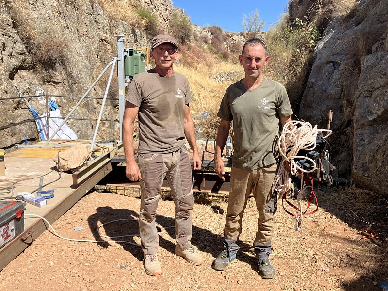 Dos técnicos de la Sociedad de Ciencias Aranzadi, vestidos con ropa de trabajo, posan en un terreno rocoso. Uno de ellos sostiene cuerdas, mientras que detrás se observa una estructura metálica con equipo.