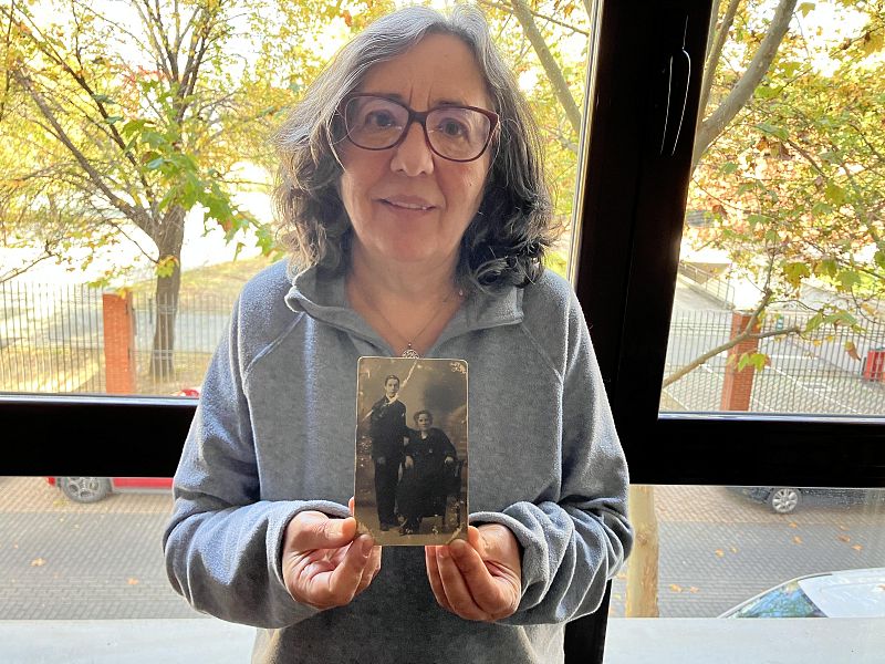 Josefa Povedano, con gafas y sudadera gris, sostiene una fotografía antigua en blanco y negro, con un hombre de pie y una mujer sentada. Se encuentra junto a una ventana con árboles de hojas amarillas.