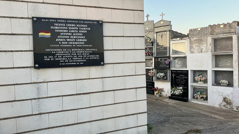 Una placa conmemorativa oscura, ubicada en una pared de ladrillo blanco, recuerda a varias personas asesinadas en Acehúche el 27 de agosto de 1936 por defender la República Española, la libertad y la democracia. La placa incluye nombres y una bandera tricolor.