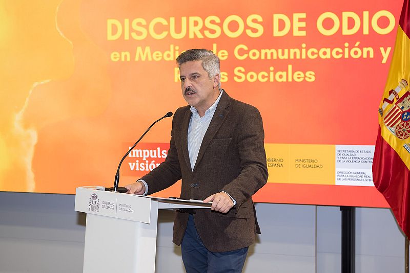 Julio del Valle, con camisa celeste y chaqueta marrón, está de pie detrás de un atril. Detrás de él, una pantalla muestra un texto y la bandera de España. El atril presenta el escudo de España y logos institucionales.
