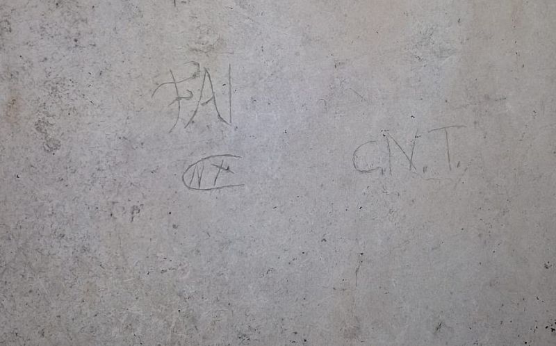 Un muro gris claro exhibe grafitis con las siglas "FAI" y "CNT", junto a un símbolo que parece una "N" dentro de un círculo con una línea, todos dibujados con trazos simples.