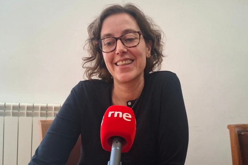 Esther Rebollar, con gafas y una blusa negra, sonríe frente a un micrófono rojo con el logo de una emisora de radio. El fondo es una pared blanca, con elementos como un radiador y una silla.