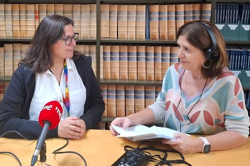 Dos mujeres, una con gafas y cinta de arcoíris frente a un micrófono, y otra con auriculares y papeles, se encuentran en una biblioteca con estanterías llenas de libros.