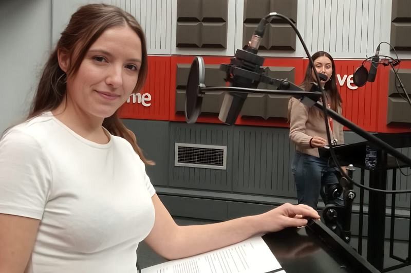En un estudio de radio, una mujer sonríe a la cámara mientras otra se prepara frente a un micrófono. El estudio cuenta con paneles acústicos y micrófonos profesionales.