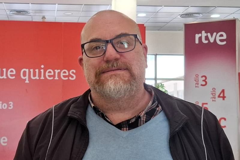 Un hombre con gafas y barba, posiblemente José María López, posa frente a un fondo con logos de RTVE y sus emisoras, vistiendo varias capas de ropa.