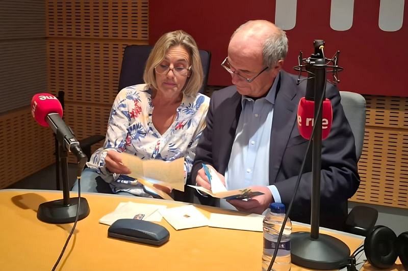 Dos personas, Adela y José Vicente Giménez Delgado, están en un estudio de radio. Adela, con gafas y blusa estampada, mira unos papeles. José Vicente, con camisa clara, escribe en una tarjeta. Hay micrófonos, una botella de agua y otros objetos sobre la mesa.